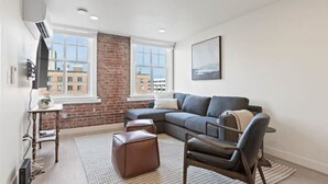 A modern living room with a sectional sofa, a leather armchair, and a small table. There are two windows with a view of buildings outside.