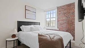 Une chambre à coucher avec un lit, une table de chevet, une lampe, une fenêtre et un mur en briques.