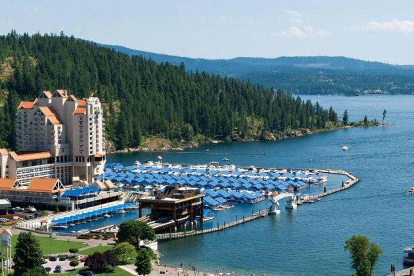 A coastal marina with multiple docks and boats, surrounded by dense forest and a large building with a red roof.