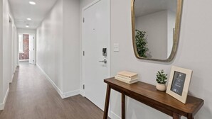 Un corridoio con una porta bianca, un tavolino consolle in legno con libri e una pianta, e uno specchio con cornice dorata.