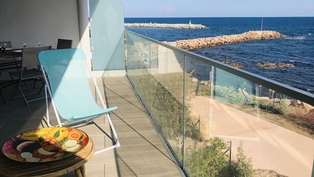 En balkon med gelænder af glas, der kigger ud over havet, et bord med en skål frugt og en stol.