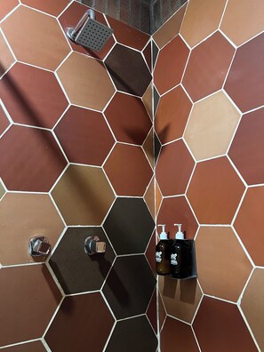 Een badkamer met honingraatmozaïek tegels in verschillende bruine en zwarte tinten, een handdouche, een zeepdispenser en een fles shampoo.