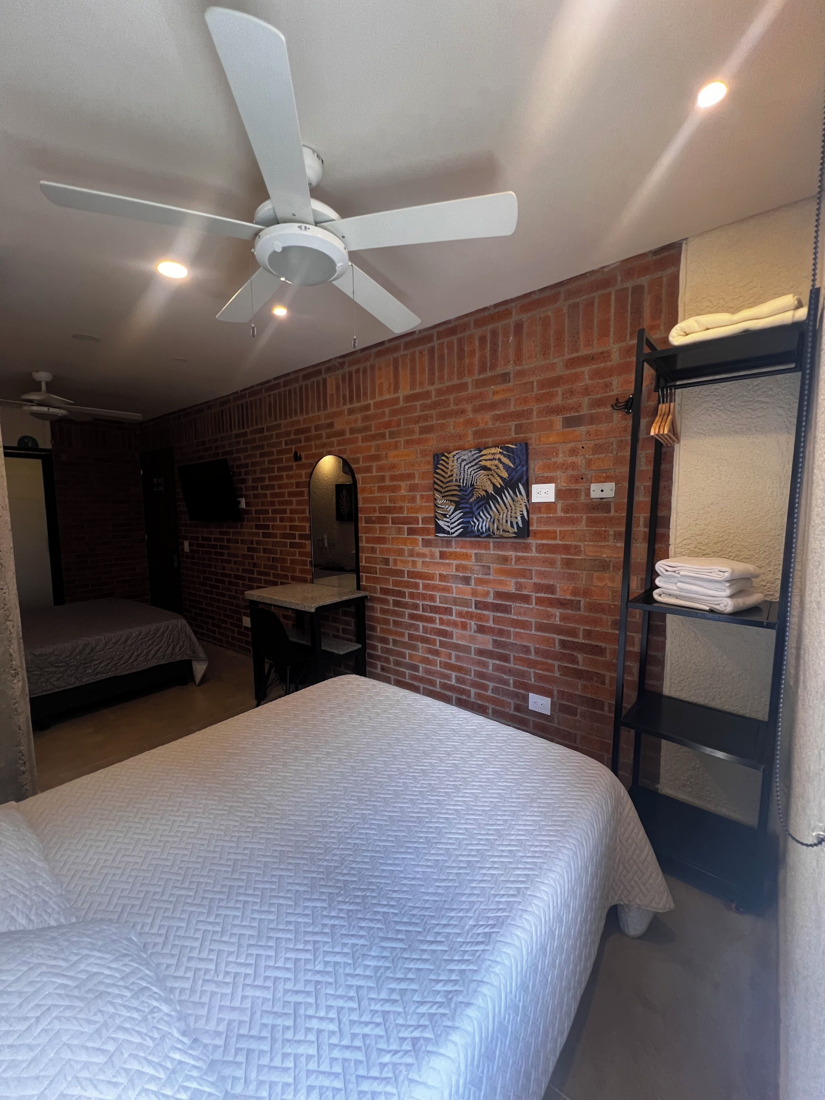 Una habitación de hotel con una cama grande, un ventilador de techo, una pared de ladrillo, un televisor de pantalla plana y un perchero.