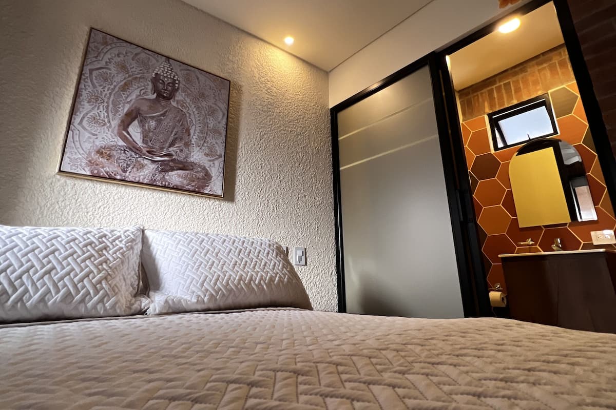 A hotel room with a bed, a ceiling fan, a wall-mounted Buddha statue, and a bathroom with a mirror and towel dispenser.