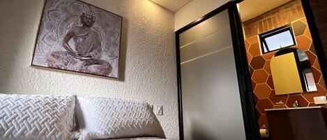 A hotel room with a bed, a ceiling fan, a wall-mounted Buddha statue, and a bathroom with a mirror and towel dispenser.