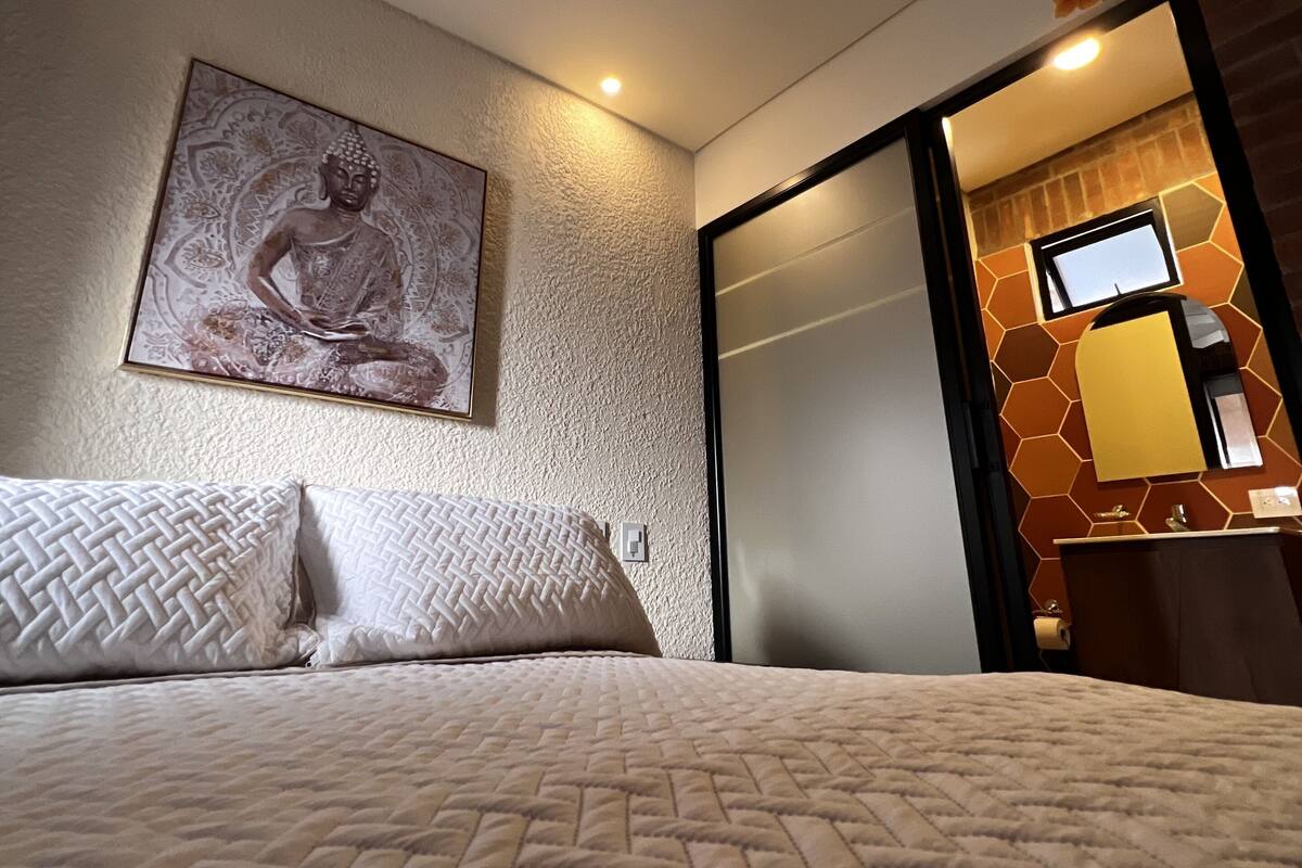 A hotel room with a bed, a ceiling fan, a wall-mounted Buddha statue, a sliding door, and a bathroom with a mirror and towel dispenser.