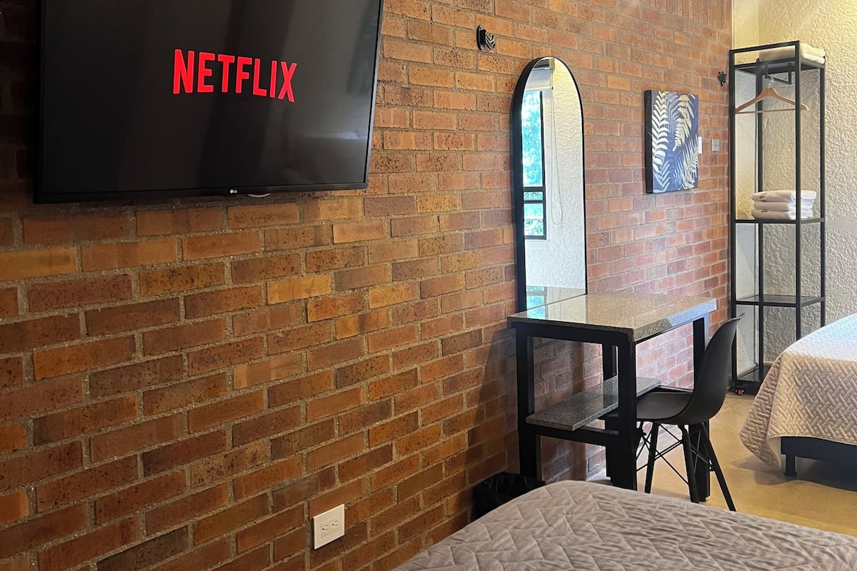 A hotel room with a brick wall, a flat-screen TV displaying the Netflix logo, a small table with a chair, a bed with a quilted cover, and a mirror.