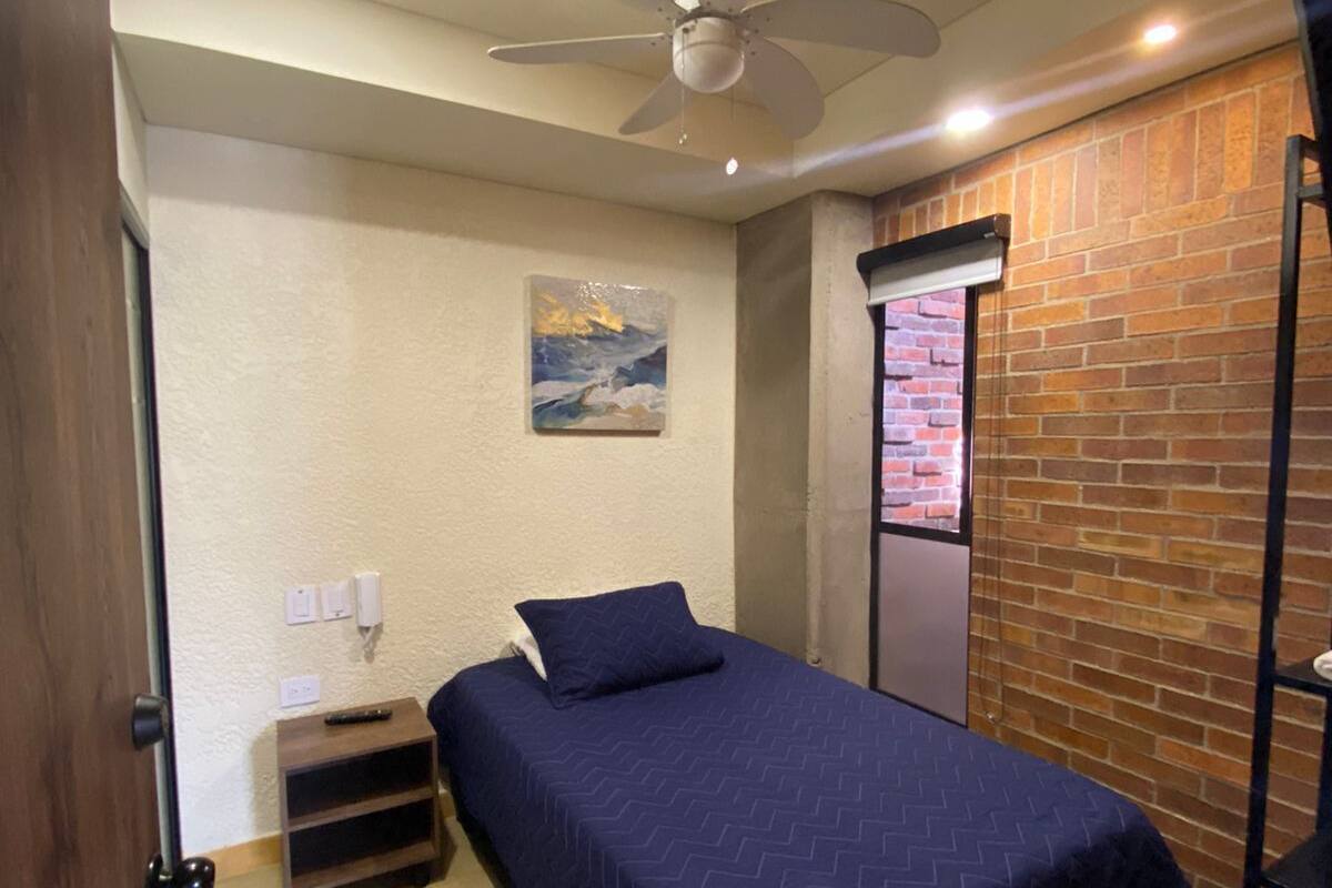 A bedroom with a bed, a ceiling fan, a painting on the wall, and a brick wall.