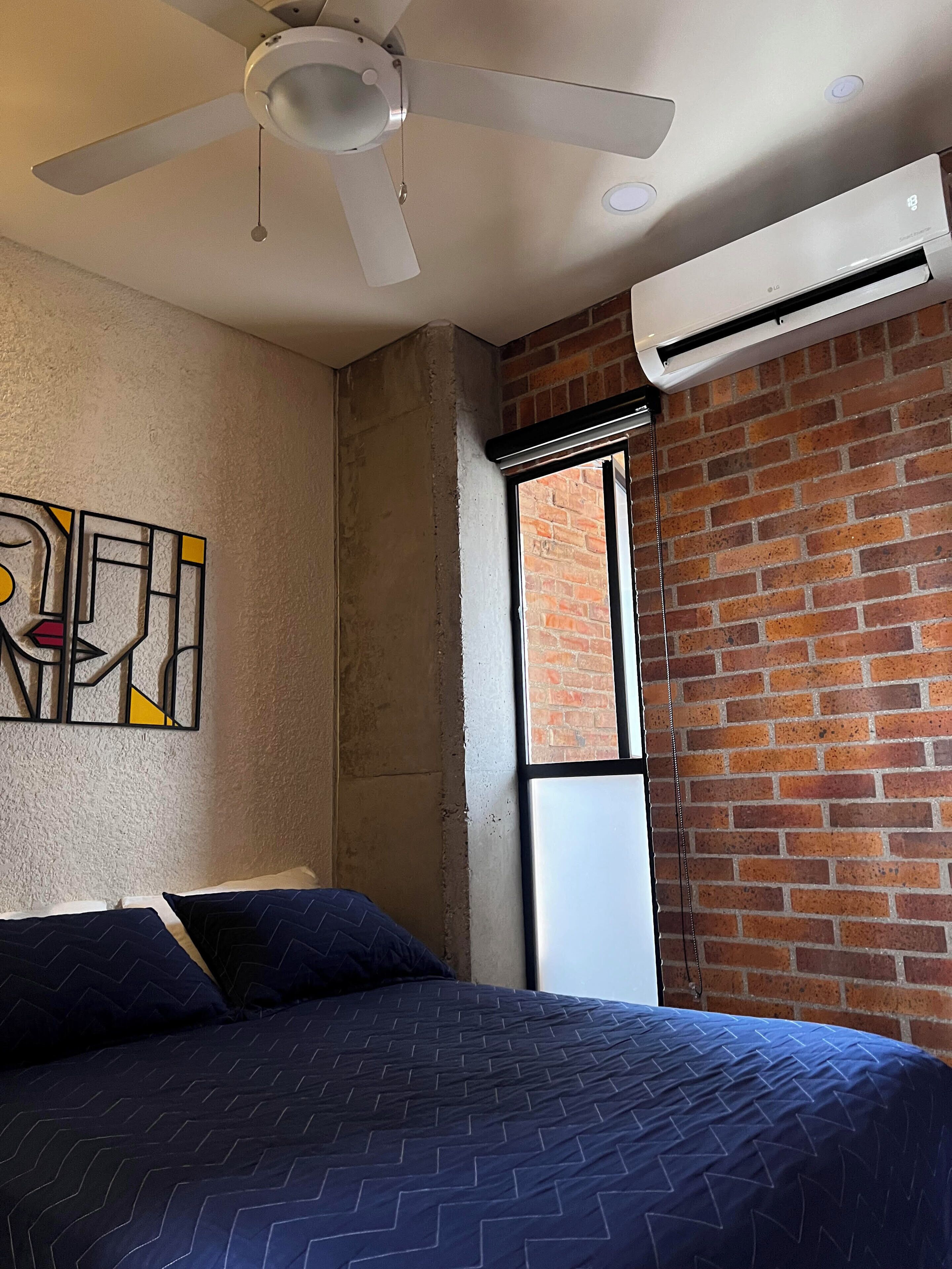 A bedroom with a bed, a ceiling fan, an air conditioning unit, a brick wall, and a window.
