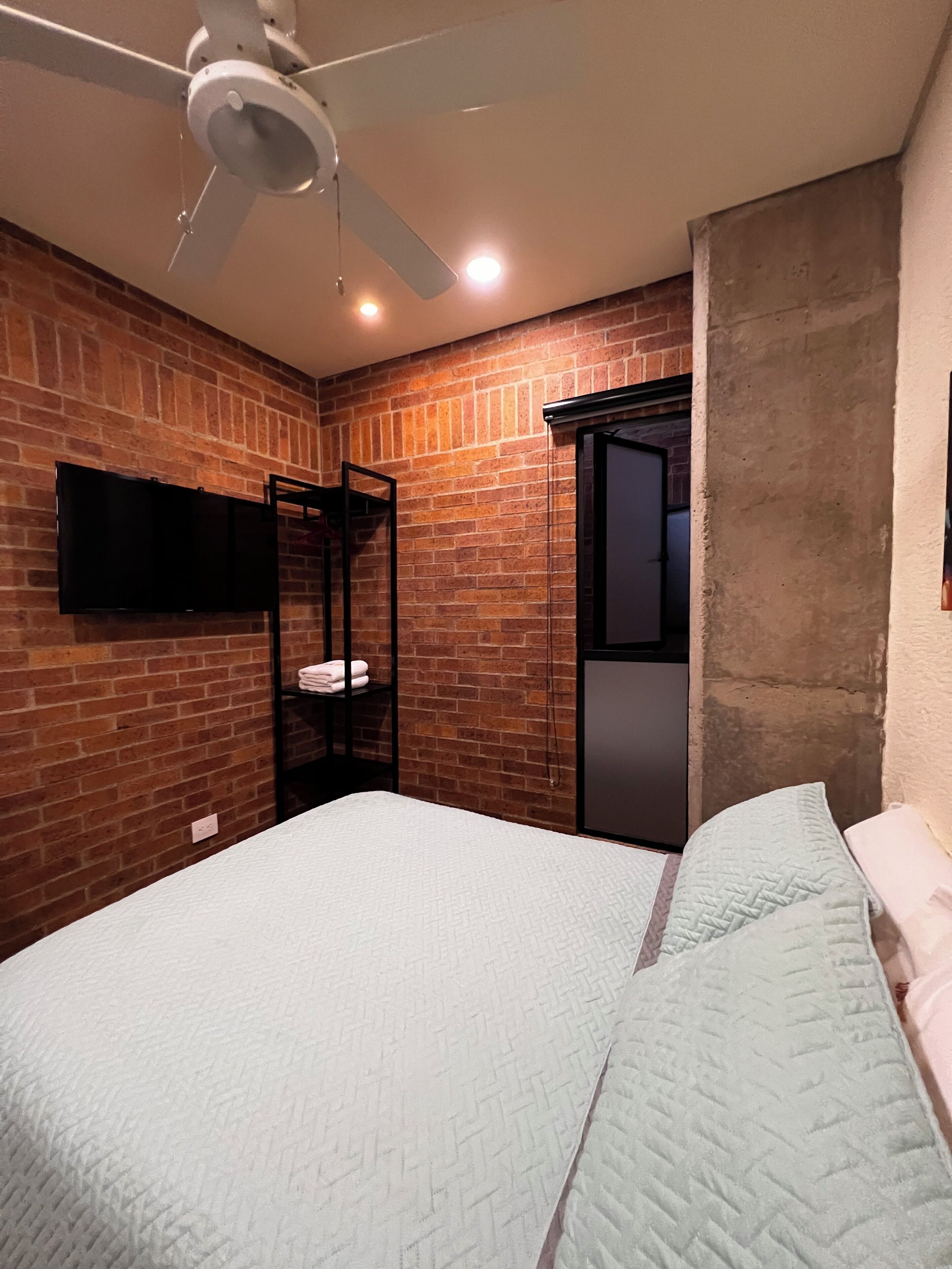 A bedroom with a brick wall, a ceiling fan, a flat-screen TV mounted on the wall, a bed with a quilted mattress, and a wardrobe.