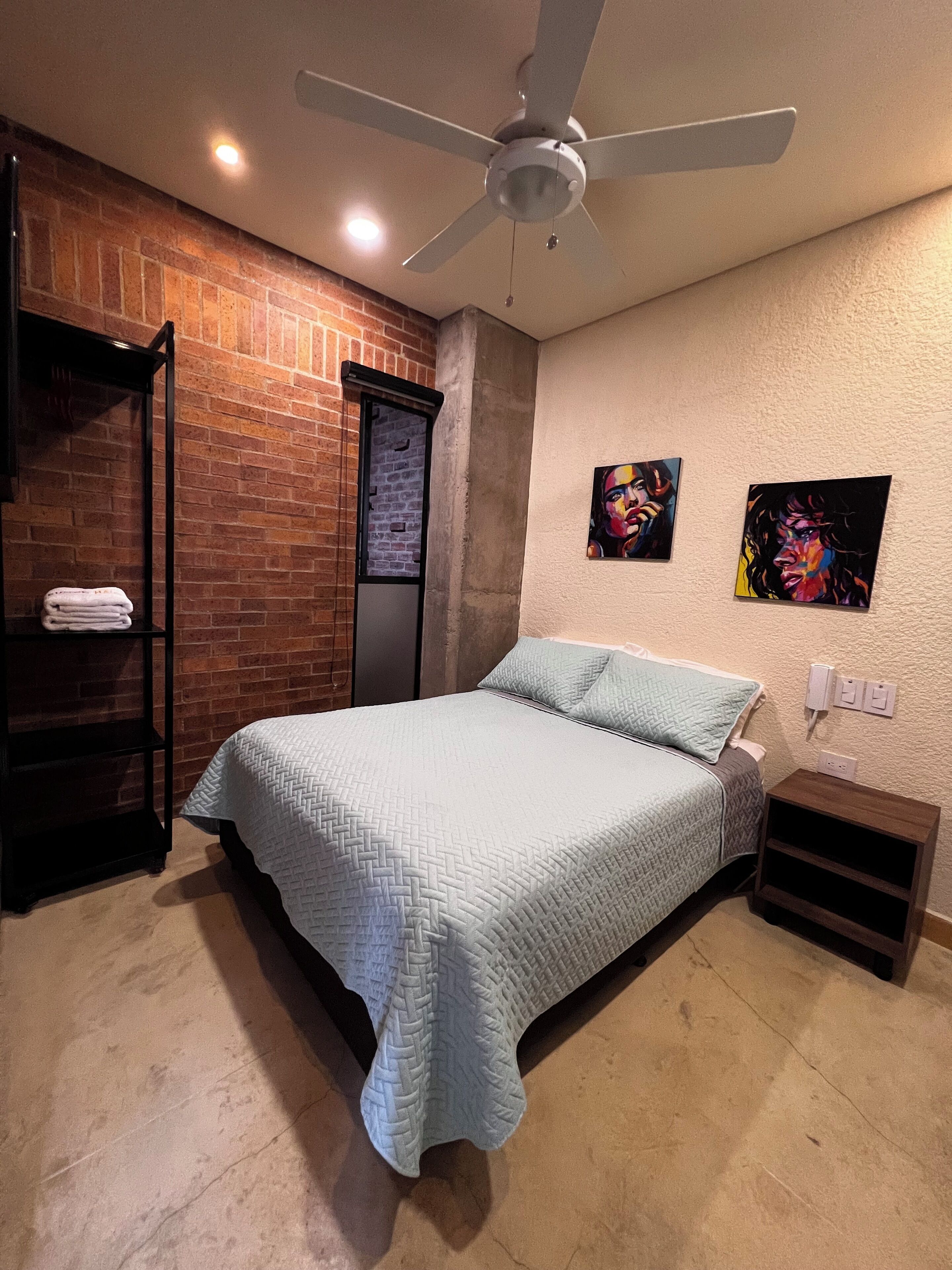 Un dormitorio con cama, ventilador de techo, pared de ladrillo y dos cuadros.