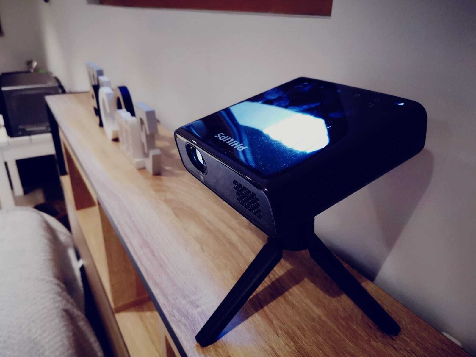 A projector on a wooden table with a white cloth, a small white object, and a wall-mounted shelf.