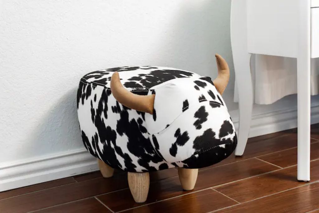 Un pouf noir et blanc en forme de vache, doté de pieds en bois, placé devant un mur blanc et sur un plancher en bois.