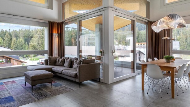 Ein modernes Wohnzimmer mit einem Ecksofa, einem Essbereich mit Holztisch und großen Fenstern mit Blick auf ein bewaldetes Gebiet.
