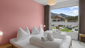 Ein Hotelzimmer mit einem großen Bett, einem Nachttisch, einer Lampe und Blick auf ein Gebäude und Berge.