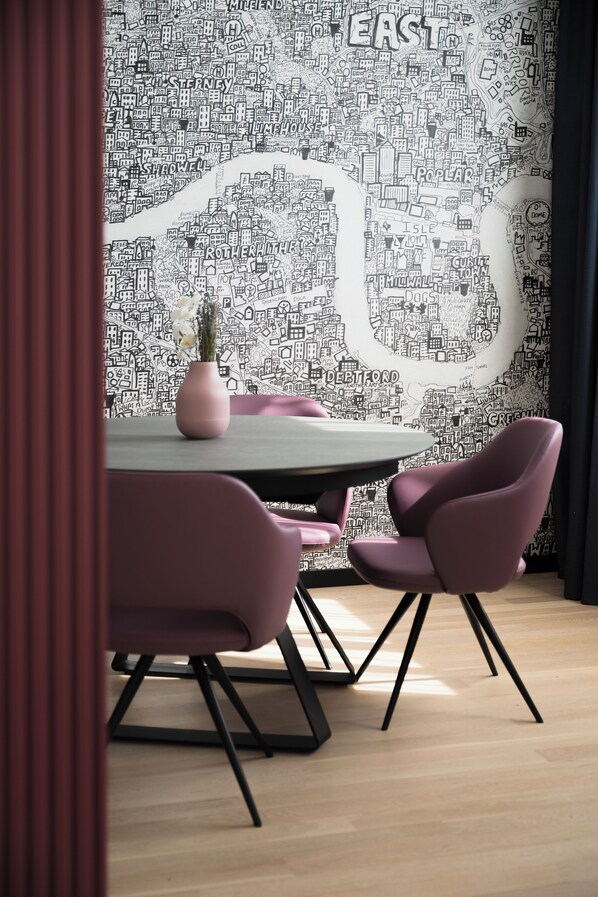 A dining area with a round table, pink chairs, and a wall with a cityscape mural. - Mitico Hotel & Natural Spa (Bologna)