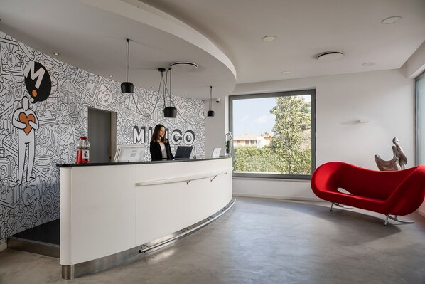 Un área de recepción moderna con un escritorio blanco, arte mural en blanco y negro, y una silla roja.