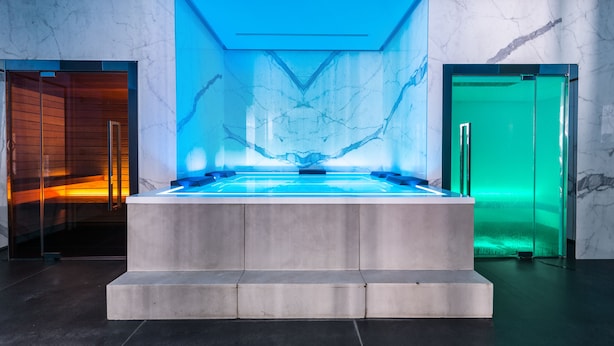 Un moderno espacio de spa con una piscina climatizada central, paredes de mármol y puertas de cristal.
