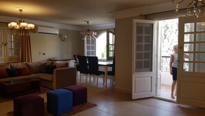 Sala de estar con sofá, mesa de centro y reposapiés. Hay un comedor con mesa y sillas. Una puerta da a un área exterior.