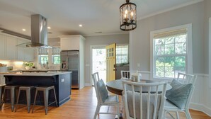 A modern kitchen with white cabinetry, a stainless steel range hood, and a central island with stools. There is a dining area with a round table and white chairs. - PREMIER Landmark Home Located in Old Town Bluffton  (Bluffton)