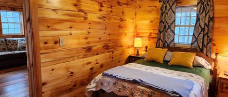 A wooden cabin bedroom with a bed, a nightstand, a lamp, and a window with curtains.