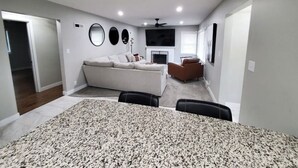 A spacious living room with a large sofa, a fireplace, and a dining area with a patterned tablecloth.