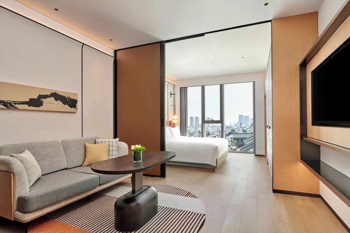 A modern hotel room with a sofa, a small table with a vase of flowers, a bed, a large TV, and a city view through large windows.