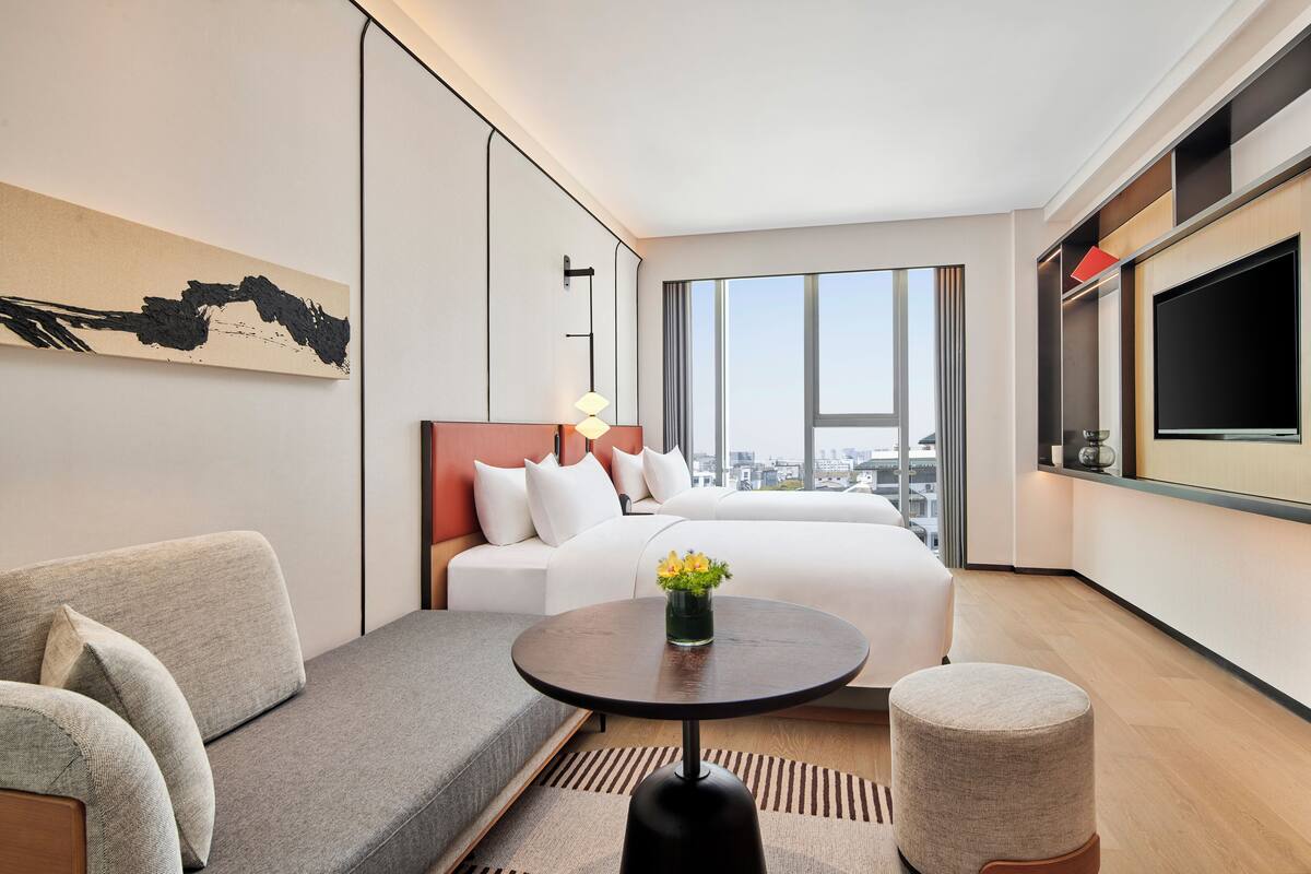 A modern hotel room with a large bed, a sofa, a small table with flowers, a TV, and a city view.