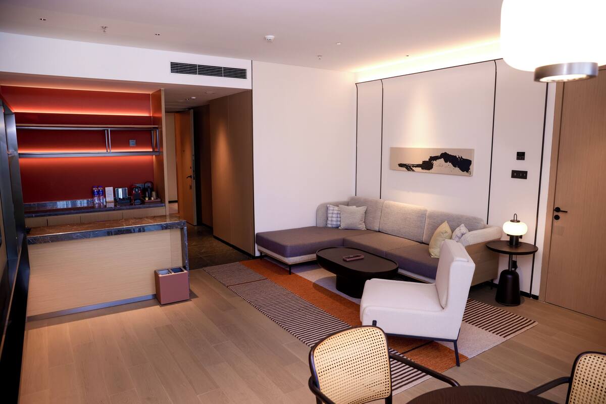 A modern hotel room with a sofa, a small table, a chair, a desk with a computer, and a lamp.