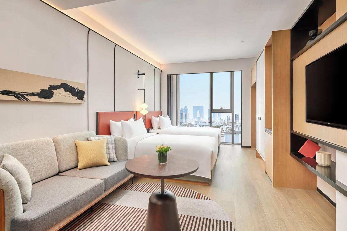 A modern hotel room with a large bed, a sofa, a TV, and a city view.