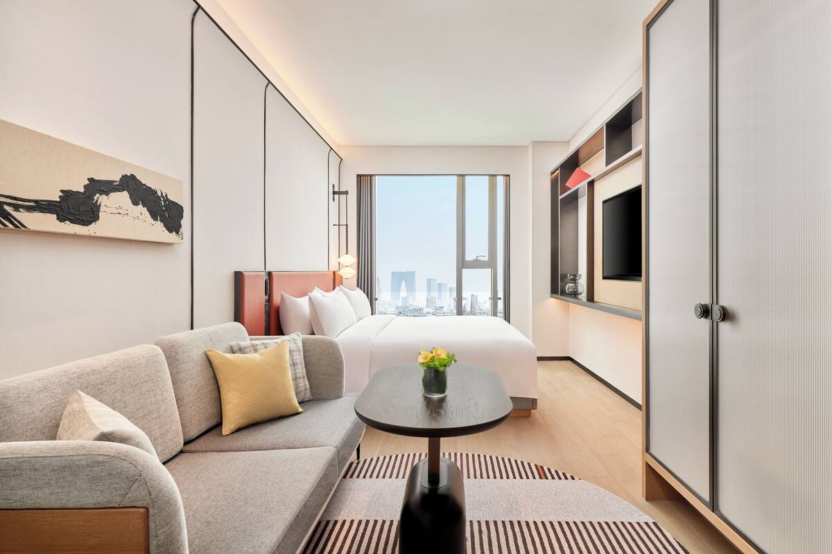 A modern hotel room with a sofa, a small table with a vase, a bed, and a wardrobe.