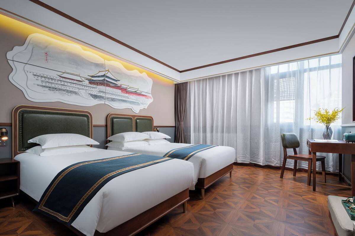 Camera d'albergo con due letti, una scrivania, una sedia, una televisione e un'ampia finestra con tende.