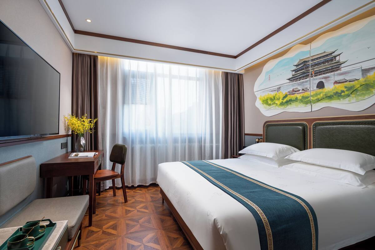 Una camera d'hotel con un letto grande, una scrivania con una sedia, un televisore e un quadro raffigurante un edificio tradizionale.