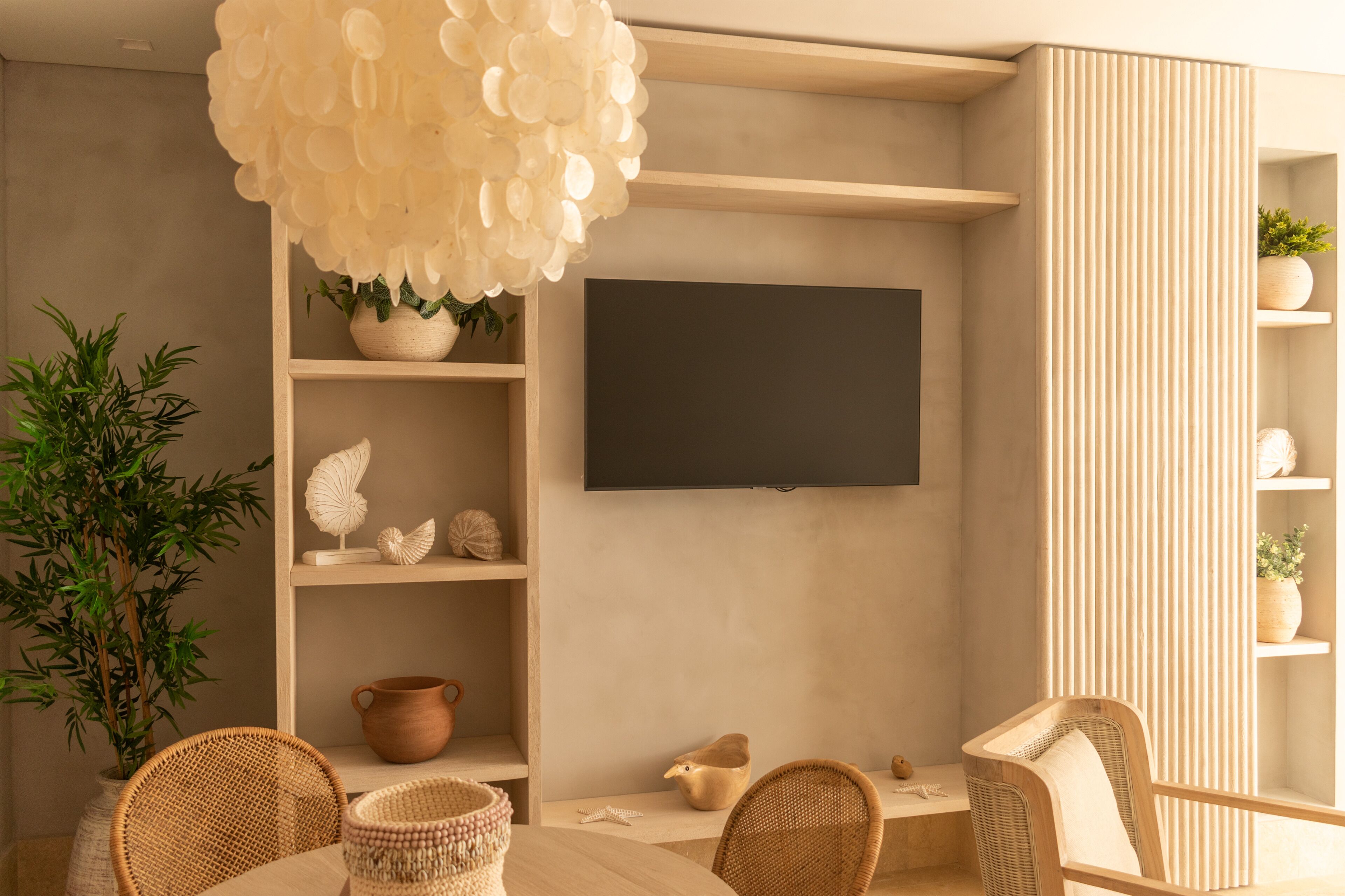 A modern living room with a flat-screen TV mounted on the wall, a large white chandelier, wicker furniture, and a potted plant.