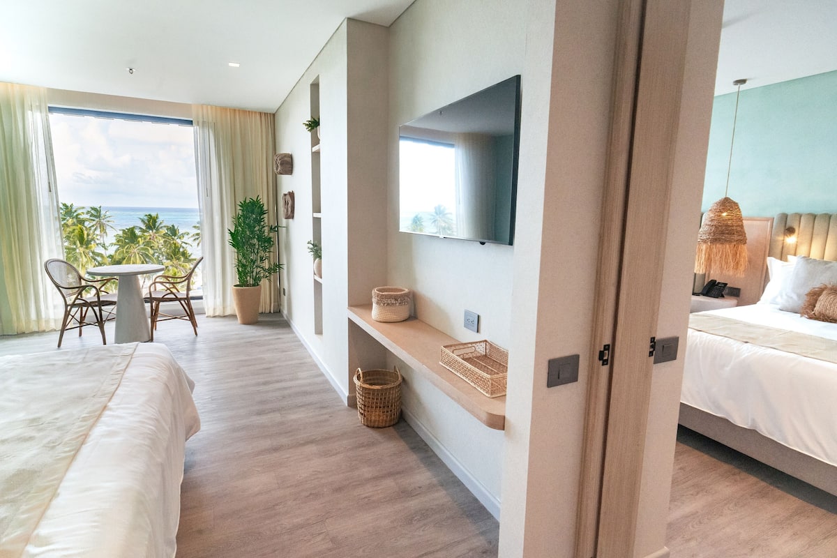 Ein modernes Hotelzimmer mit einem großen Bett, einem an der Wand befestigten Fernseher, einem Balkon mit Meerblick und einer kleinen Sitzecke mit Tisch und Stühlen.