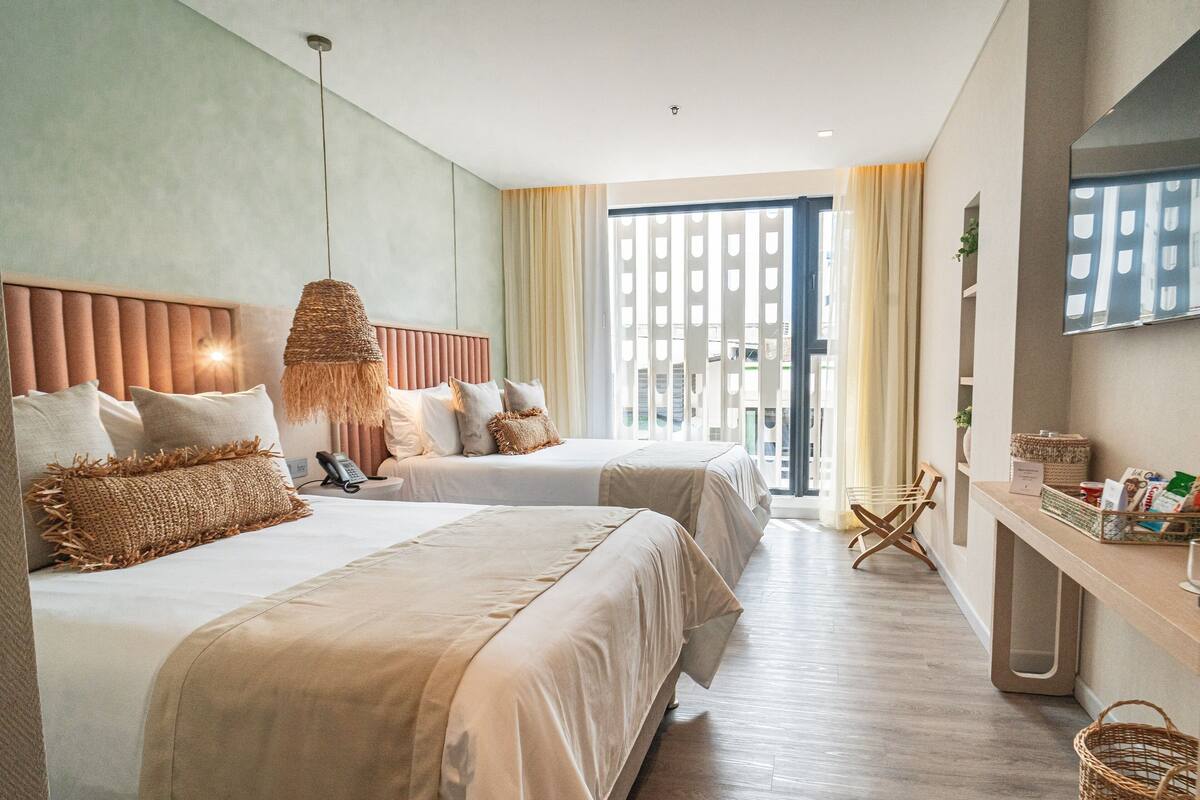 Ein Hotelzimmer mit zwei Betten, einem Schreibtisch mit Korb, einer Hängelampe und einem großen Fenster mit Vorhängen.