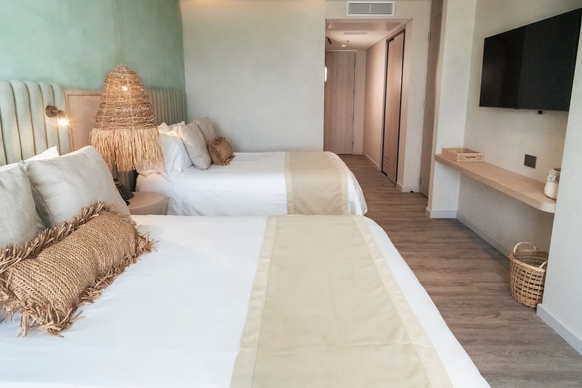 Ein Hotelzimmer mit zwei Betten, einer gewebten Lampe, einem an der Wand befestigten Fernseher und einem Holzboden.