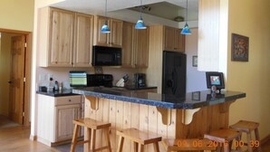 Une cuisine avec des meubles de rangement en bois, un réfrigérateur noir, un four à micro-ondes et un plan de travail bleu. On y trouve trois lampes suspendues et deux tabourets en bois.