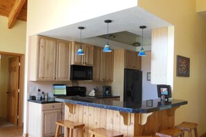 A kitchen with wooden cabinets, a black refrigerator, a microwave, and a blue countertop. There are three hanging pendant lights and two wooden stools.