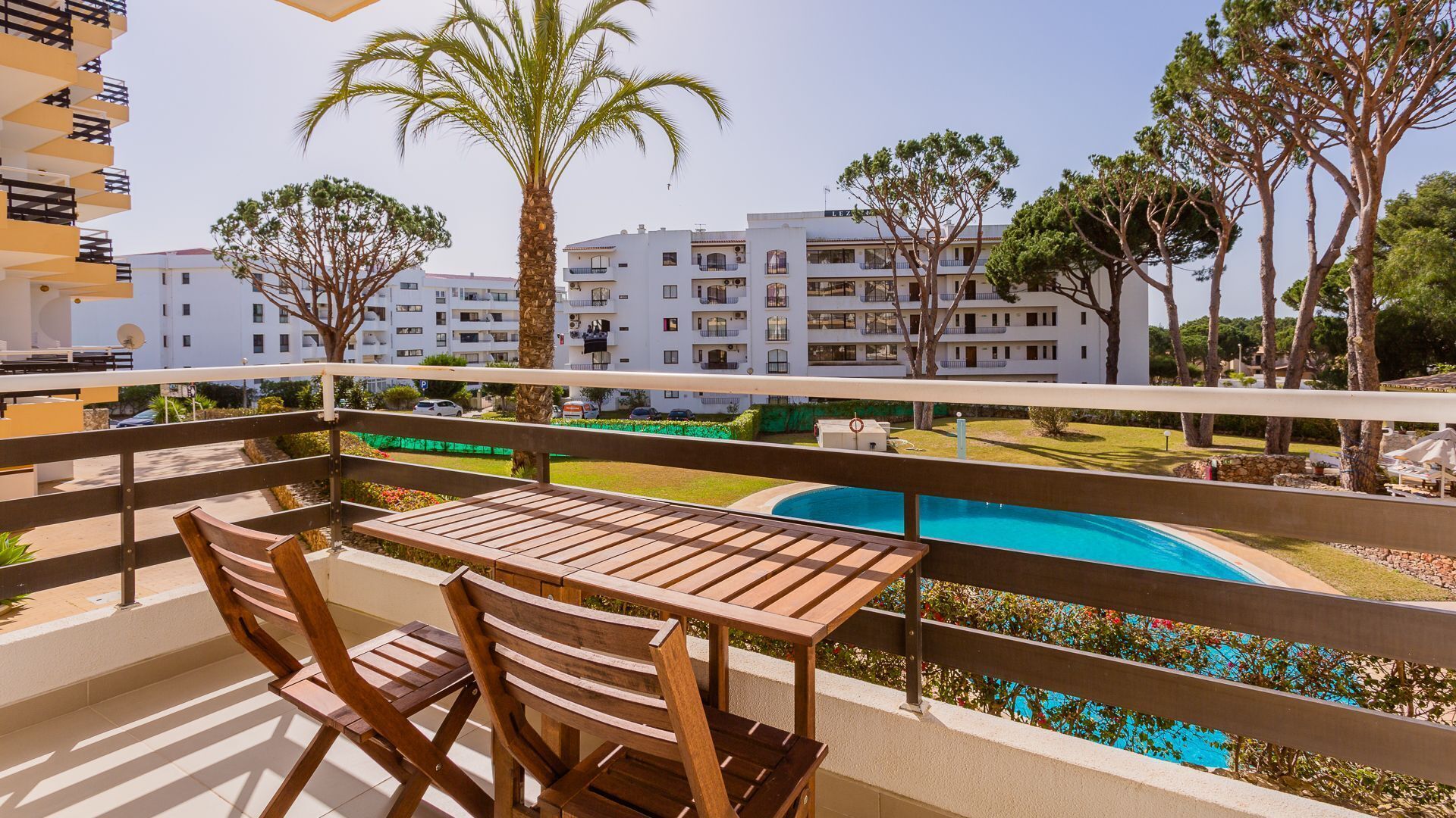 A balcony with wooden furniture overlooking a swimming pool and a residential area with white buildings and palm trees.