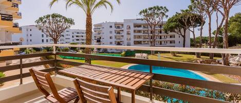 A balcony with wooden furniture overlooking a swimming pool and a residential area with white buildings and palm trees.