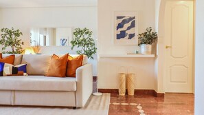 A living room with a sofa, cushions, a coffee table, a rug, a plant, a wall-mounted shelf with decorative items, and a door.