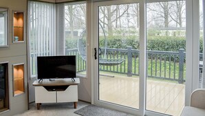 Un salon moderne avec un téléviseur à écran plat posé sur un meuble blanc, de grandes portes-fenêtres coulissantes donnant sur une terrasse avec une plateforme en bois et une clôture en métal.