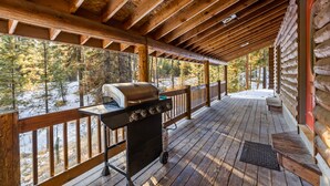 A covered wooden deck with a grill and a view of a snowy forest.