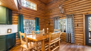 A log cabin interior with a dining table, chairs, a clock, and a deer head decoration.