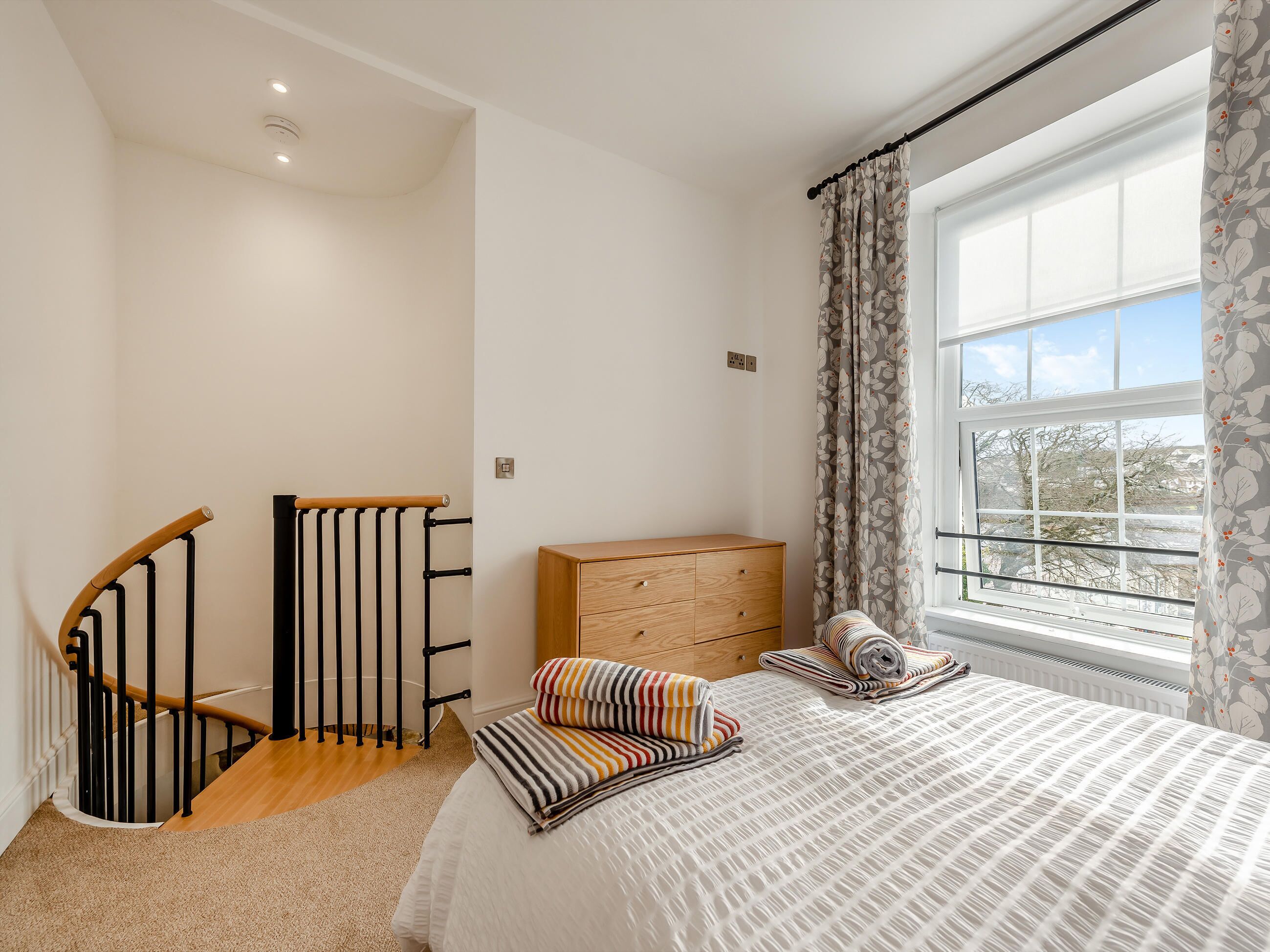 Une chambre à coucher comprenant un lit, une commode en bois, une fenêtre avec des rideaux, un escalier avec une rampe et un radiateur.