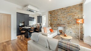 Un salon moderne avec une cheminée en pierre, un coin repas avec des tabourets de bar et une cuisine avec des meubles de couleur sombre.