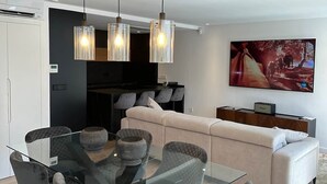 A modern living room with a glass dining table, grey upholstered chairs, a beige sofa, and a large abstract painting on the wall.