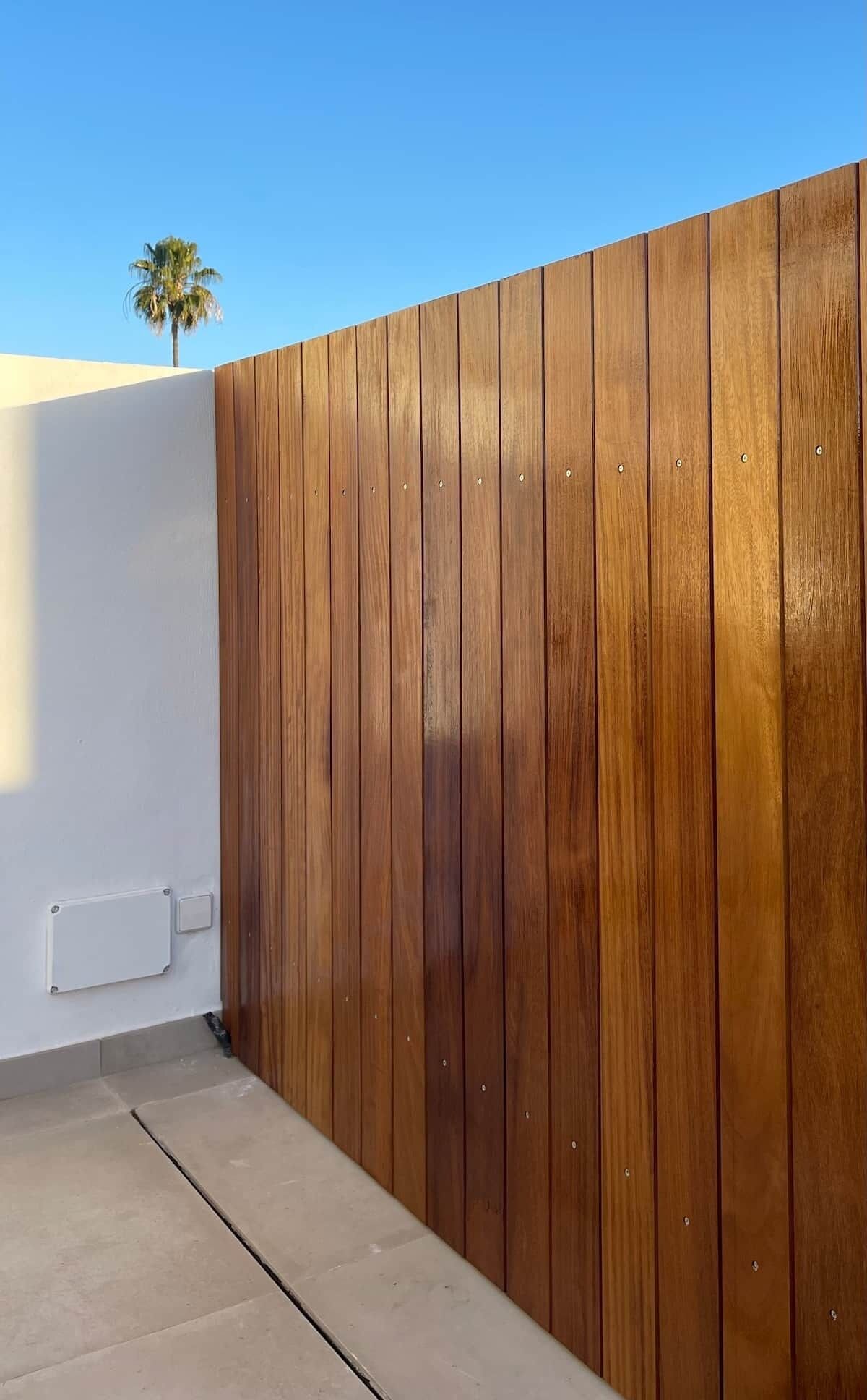 A wooden fence with a white wall and a palm tree in the background.