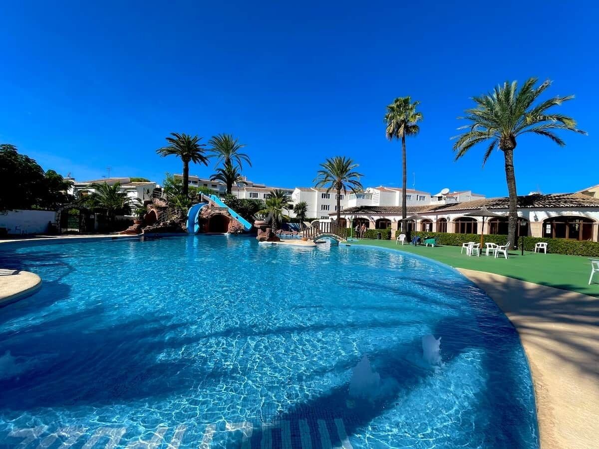 Una gran piscina con tobogán de agua, palmeras y un edificio con techo de tejas al fondo.